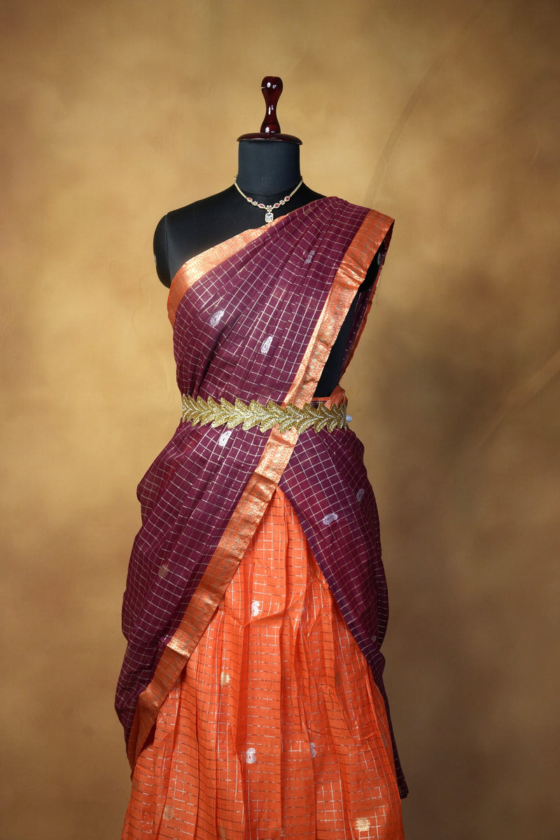 A traditional Indian saree displayed on a black mannequin torso against a warm brown studio backdrop. The saree has a deep purple upper drape with a subtle grid pattern, a metallic gold waist belt, and a bright orange lower drape with small white motifs.