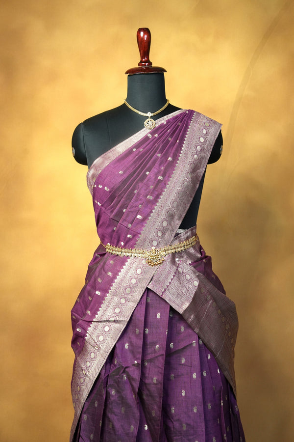 A purple and lavender saree with silver zari motifs, draped on a black mannequin torso against a warm golden-yellow studio background.