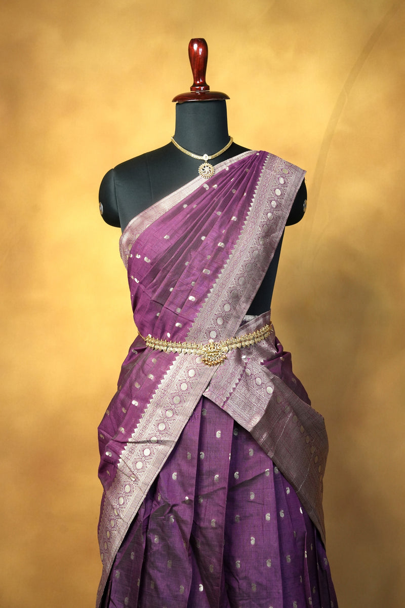 A purple and lavender saree with silver zari motifs, draped on a black mannequin torso against a warm golden-yellow studio background.