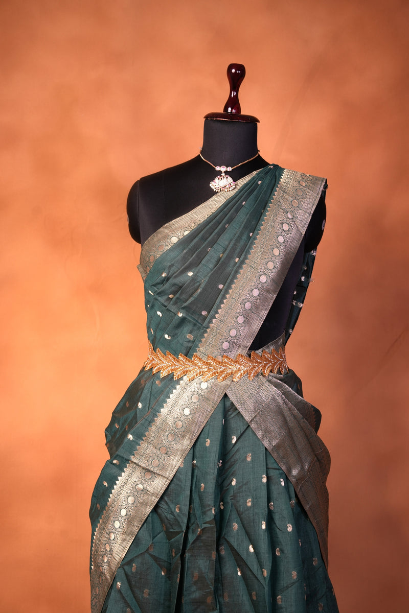 A dark teal green saree with silver zari embroidery and a wide silver border, draped on a black mannequin torso against a warm orange-brown studio background.