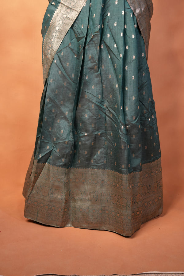 A dark teal green saree with small gold motifs and a wide brown border, draped against a solid terracotta-colored background.