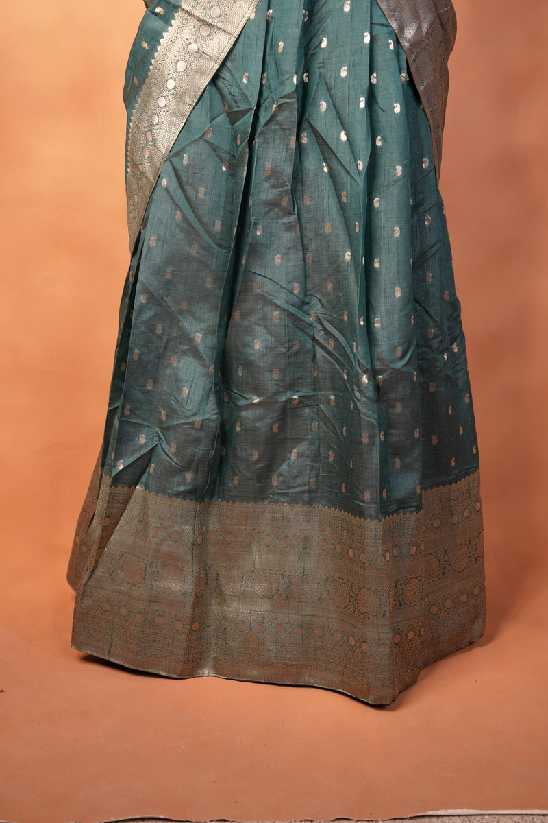 A dark teal green saree with small gold motifs and a wide brown border, draped against a solid terracotta-colored background.