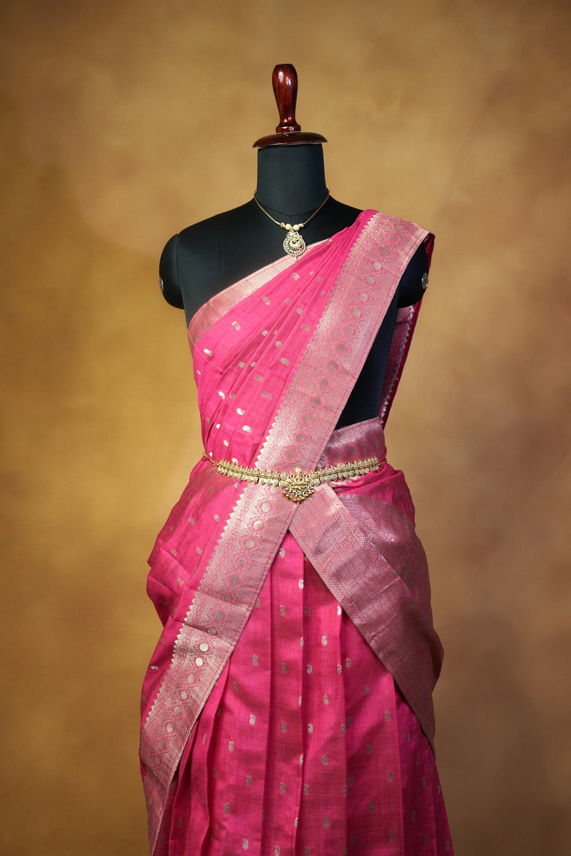 A pink silk saree with a metallic gold border and small gold motifs, draped on a black mannequin torso against a warm brown textured background.