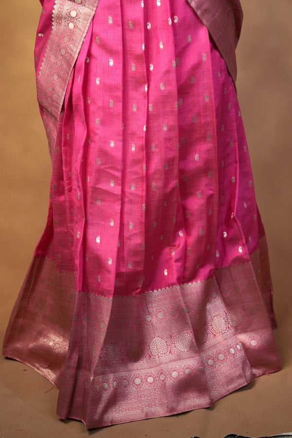 A close-up of a pink silk saree with metallic silver zari motifs and a wide decorative border, draped against a plain tan background.