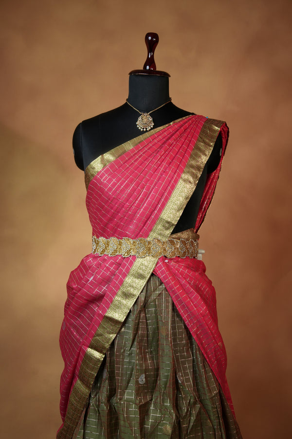 A draped sari displayed on a black mannequin torso against a warm brown studio background. The fabric is predominantly pink with a metallic gold border and a secondary olive green section with gold woven detailing.