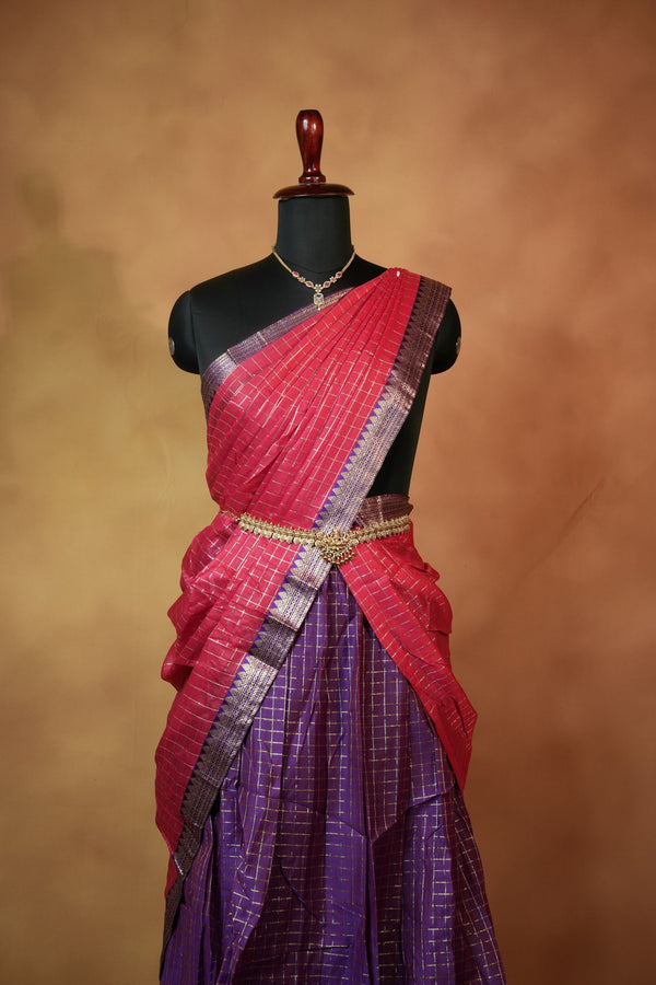 A traditional Indian saree displayed on a black mannequin torso against a warm brown studio backdrop. The saree has a vibrant pink upper drape with a metallic silver border, a gold waist belt, and a purple lower drape with a subtle grid pattern.
