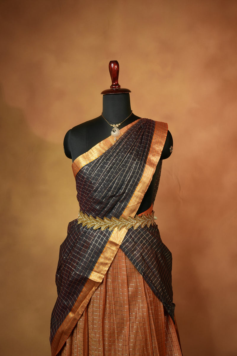 A dark brown and black checkered saree draped on a black mannequin torso, featuring a gold embroidered waist detail and a metallic gold border, set against a warm brown textured background.