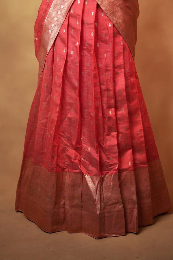 A close-up of a pleated red fabric garment, likely a skirt or drape, with a metallic gold border and small gold motifs, set against a solid tan background.