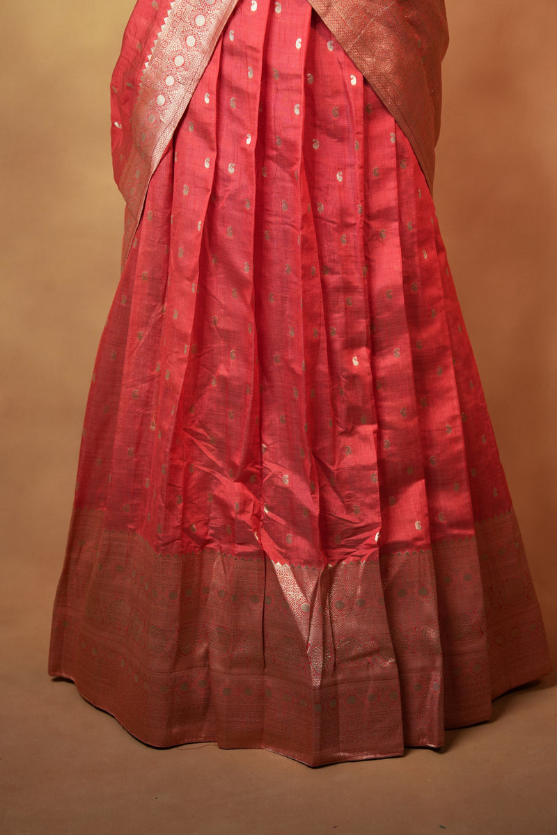 A close-up of a pleated red fabric garment, likely a skirt or drape, with a metallic gold border and small gold motifs, set against a solid tan background.