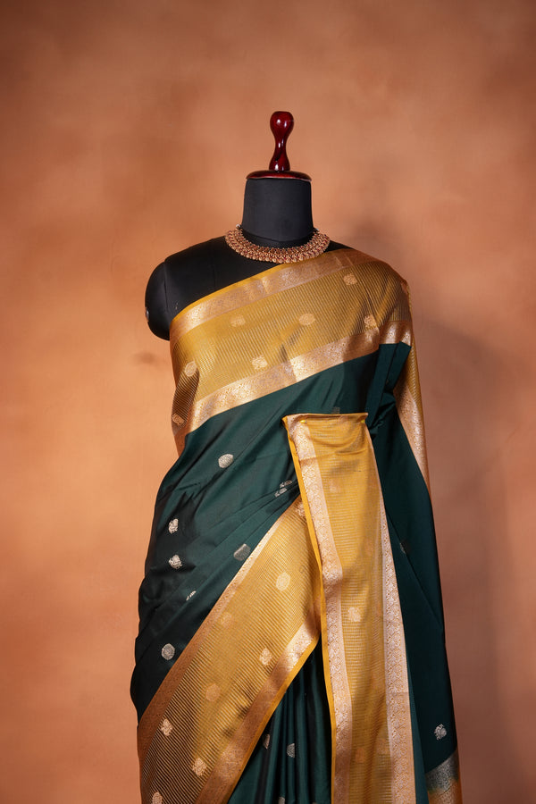 A dark green and gold saree draped elegantly on a black dress form mannequin, set against a warm, textured brown background.