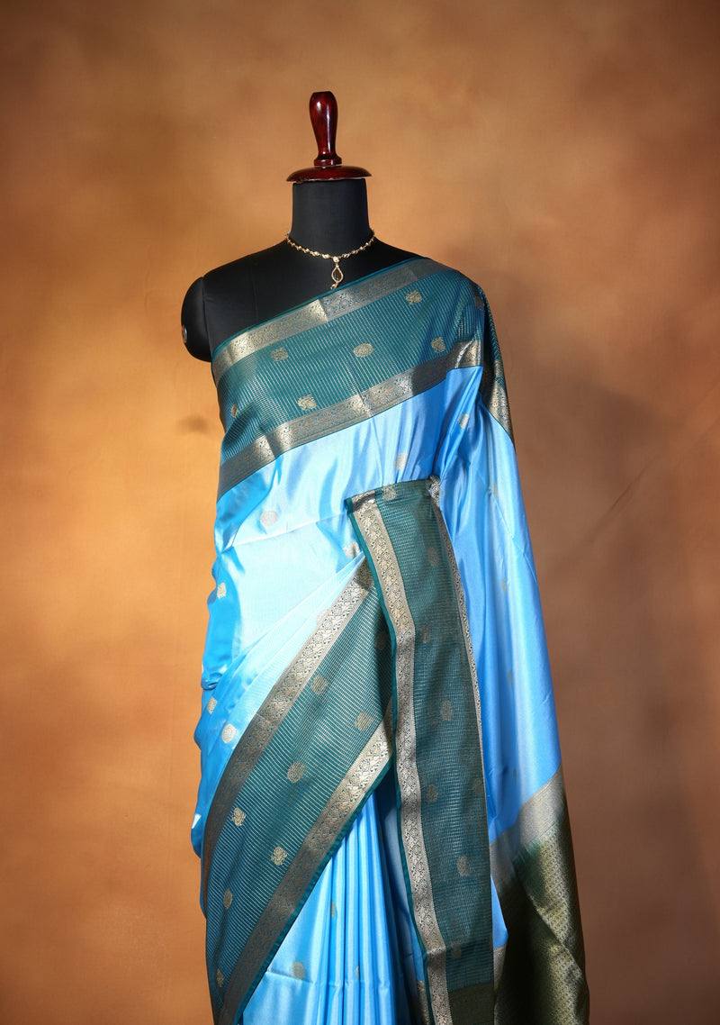 A light blue and dark teal silk saree with metallic gold zari borders, displayed on a black mannequin torso against a warm brown studio background.