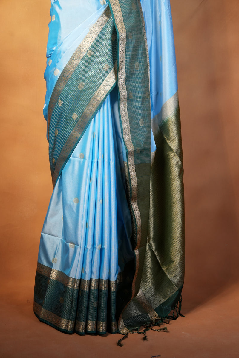 A folded light blue saree with a dark teal border and metallic gold zari patterns, draped against a warm brown background.