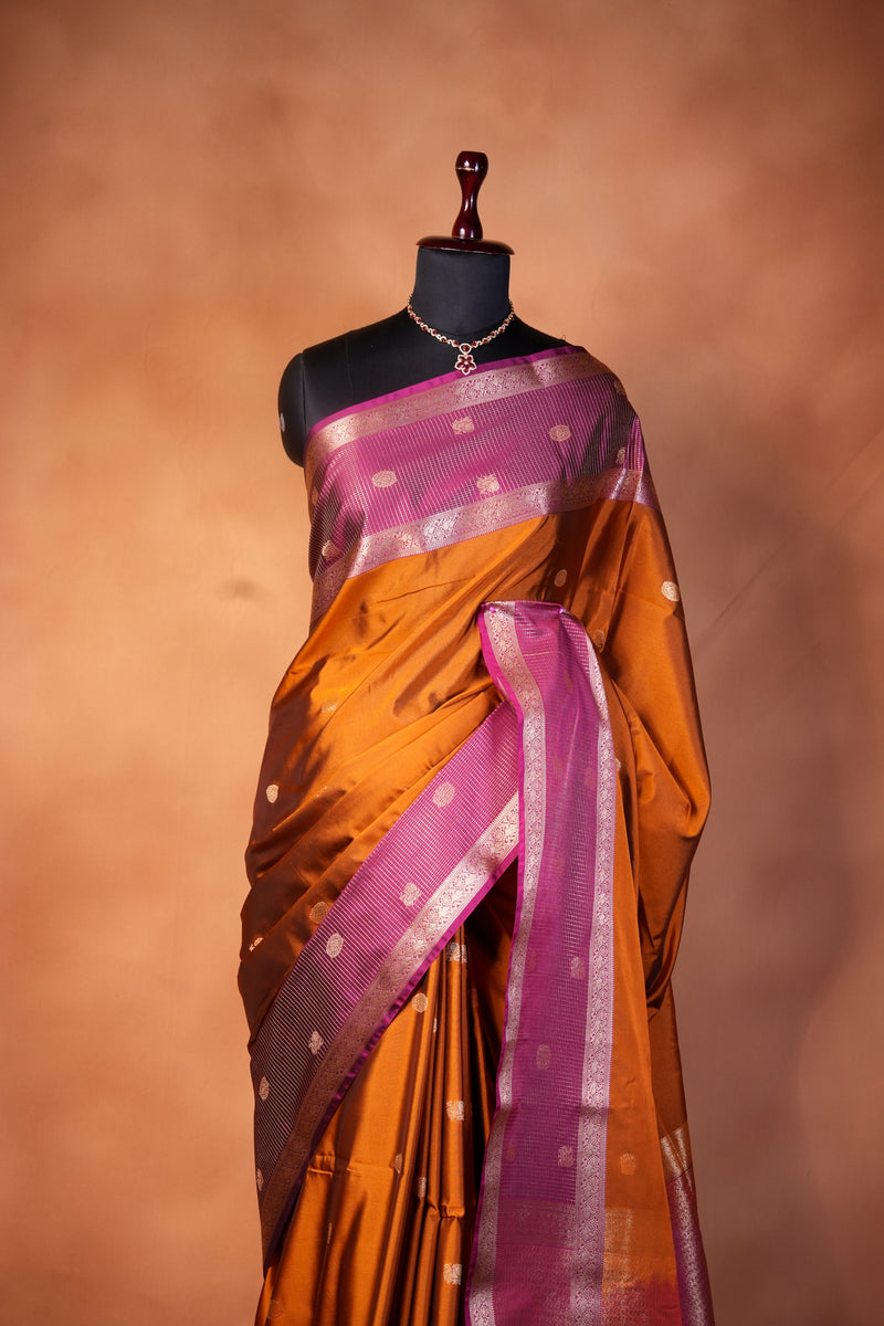 An orange and purple silk saree draped elegantly on a black mannequin torso, set against a warm, solid brown background.