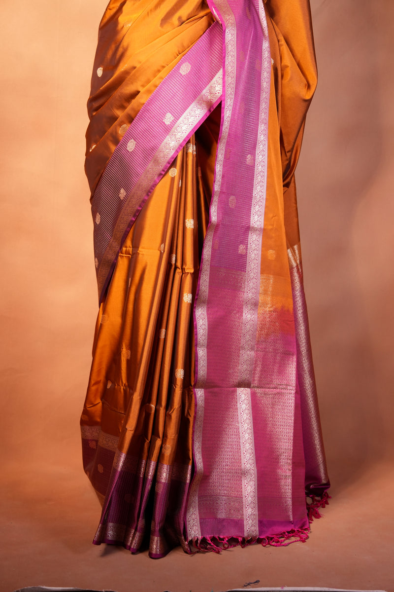 A folded and draped silk saree with a metallic bronze-gold body, a vibrant magenta-pink border, and silver zari woven patterns, set against a solid warm brown background.