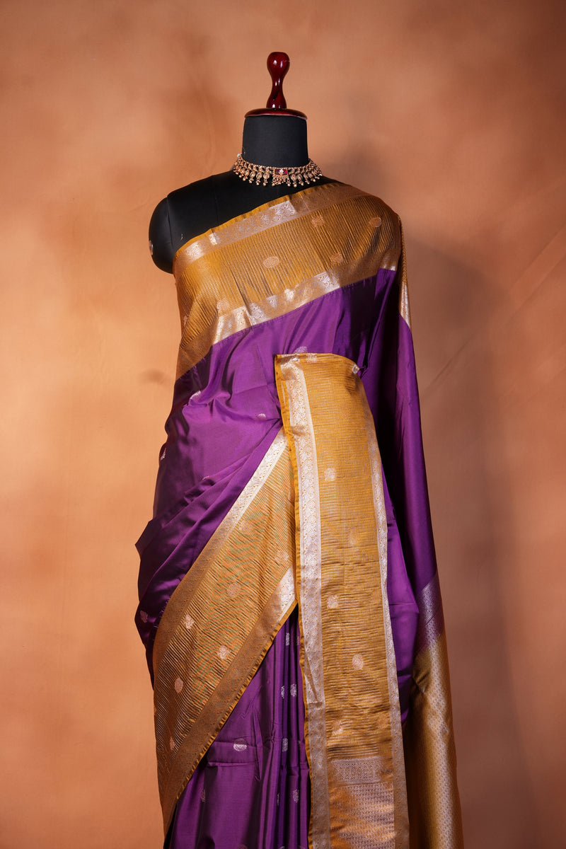 A close-up of a draped purple and gold fabric, likely a sari, displayed on a black dress form mannequin against a warm, solid brown background.