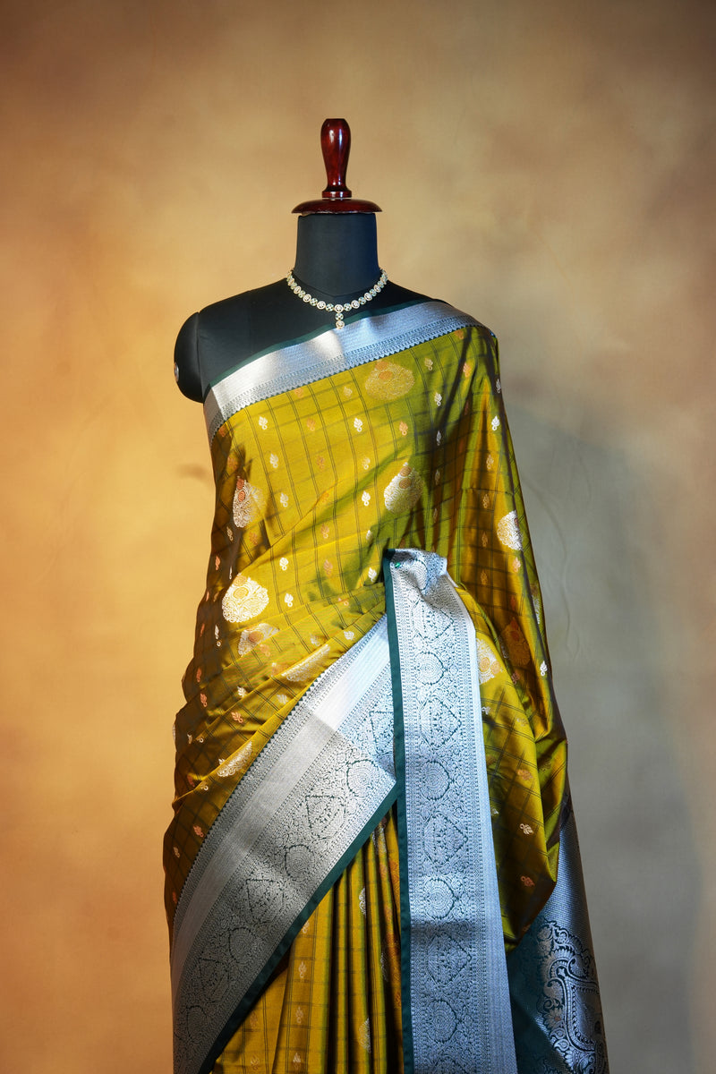 A yellow and silver Banarasi silk saree with gold zari motifs, draped on a black mannequin torso against a warm brown gradient background.