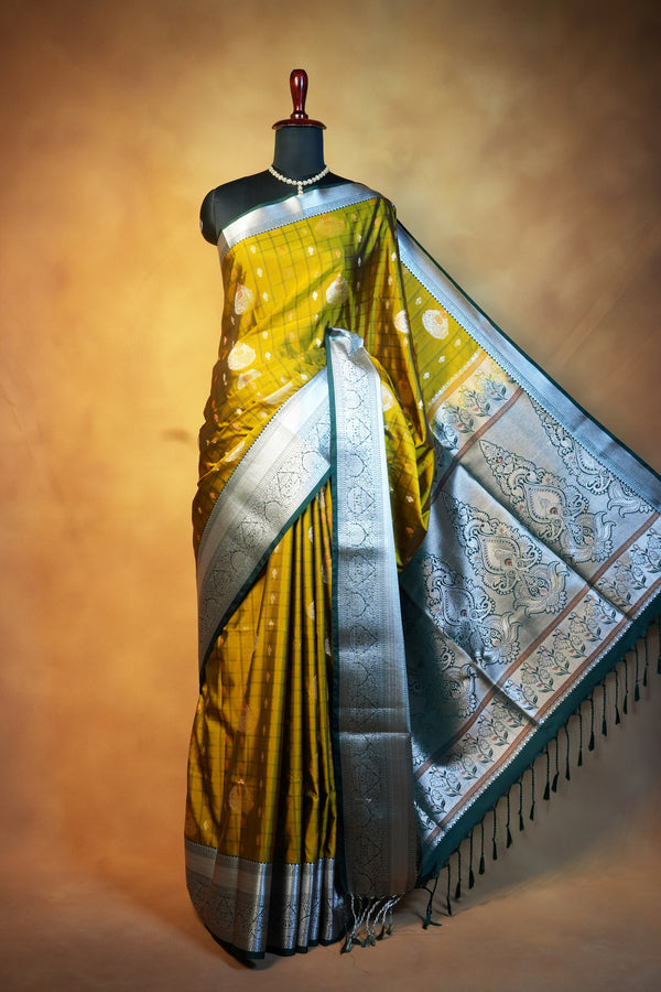 A yellow and silver Banarasi silk saree displayed on a black mannequin torso against a warm brown studio backdrop. The saree features a rich yellow body with metallic silver borders, intricate silver zari embroidery, and a matching silver patterned pallu with fringe.