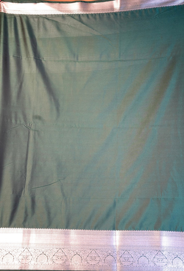 A dark teal green fabric, likely a saree, shown hanging flat. It features a wide, decorative silver border with intricate geometric and floral patterns along the bottom edge.