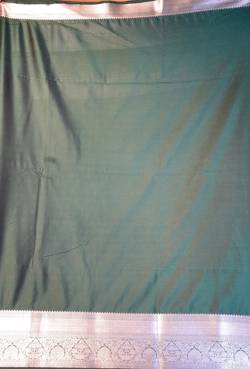 A dark teal green fabric, likely a saree, shown hanging flat. It features a wide, decorative silver border with intricate geometric and floral patterns along the bottom edge.