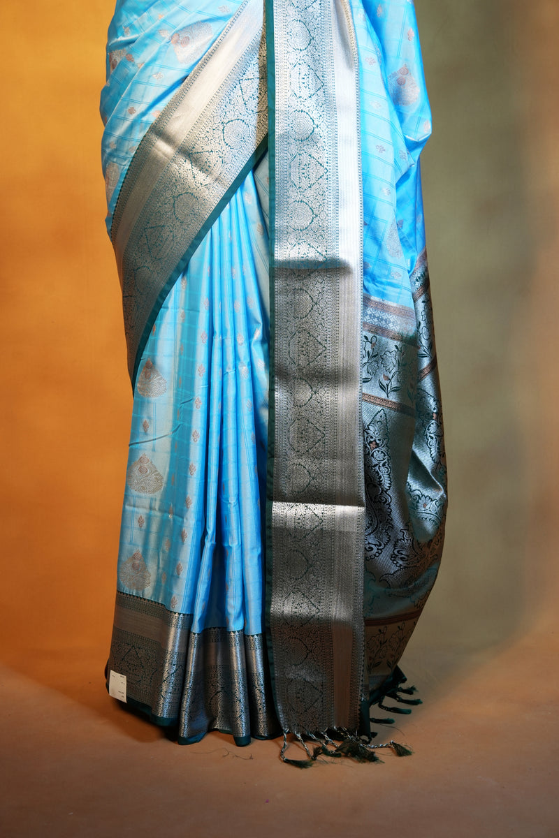A close-up of a light blue silk saree with a wide metallic silver border and intricate woven patterns, draped on a model against a warm brown background.