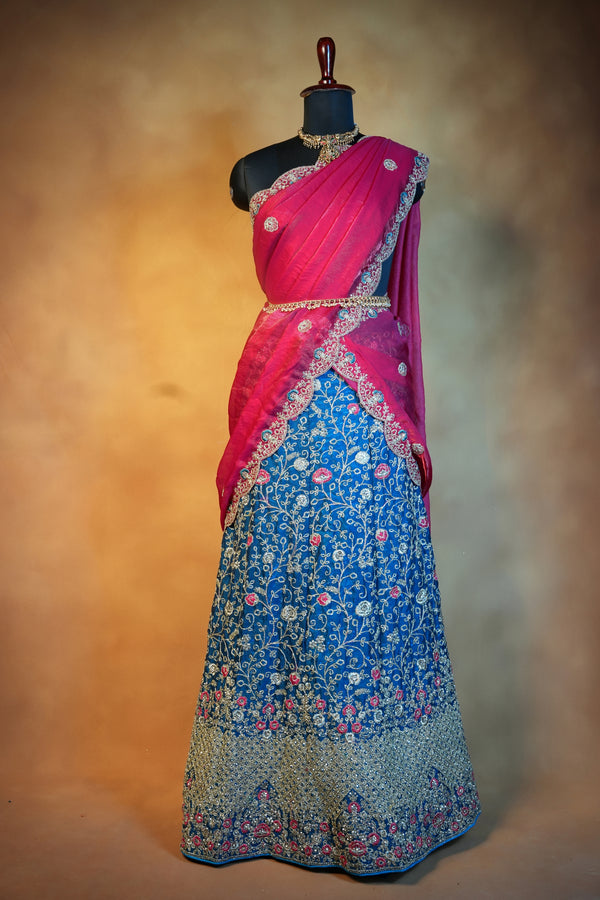A traditional Indian lehenga choli set displayed on a black mannequin. The outfit features a bright pink sleeveless blouse with silver embroidery, a matching pink dupatta, and a blue embroidered skirt with floral patterns and metallic borders.