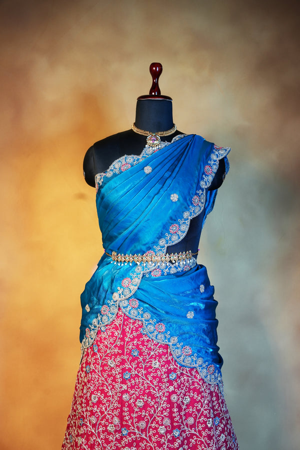 A traditional Indian outfit displayed on a black mannequin, featuring a bright blue sari draped over a pink embroidered skirt with intricate silver and gold floral patterns.