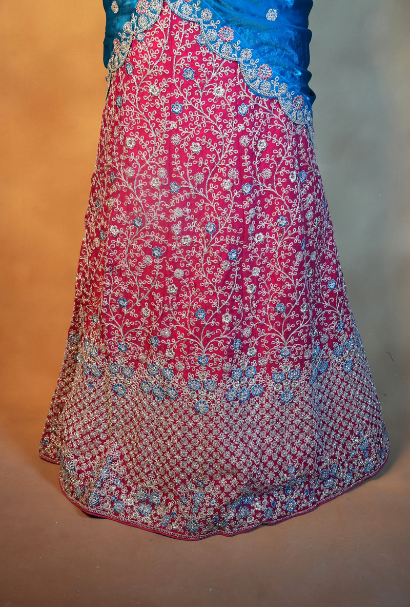 A close-up of a traditional Indian lehenga skirt in vibrant pink with intricate silver and blue floral embroidery, paired with a blue embroidered blouse top, set against a plain beige background.