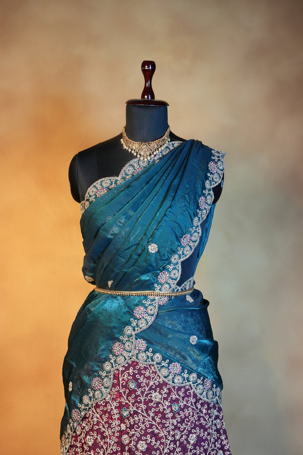 A teal blue embroidered sari draped on a black dress form mannequin, featuring intricate silver floral embroidery and a contrasting magenta embroidered section on the left, set against a warm beige background.