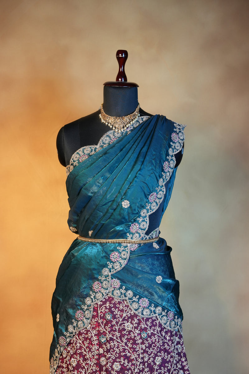 A teal blue embroidered sari draped on a black dress form mannequin, featuring intricate silver floral embroidery and a contrasting magenta embroidered section on the left, set against a warm beige background.