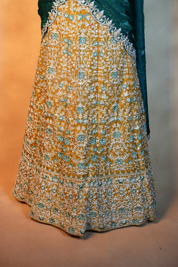 A close-up of a traditional embroidered skirt in mustard yellow with intricate white and teal floral embroidery, paired with a dark teal draped fabric.