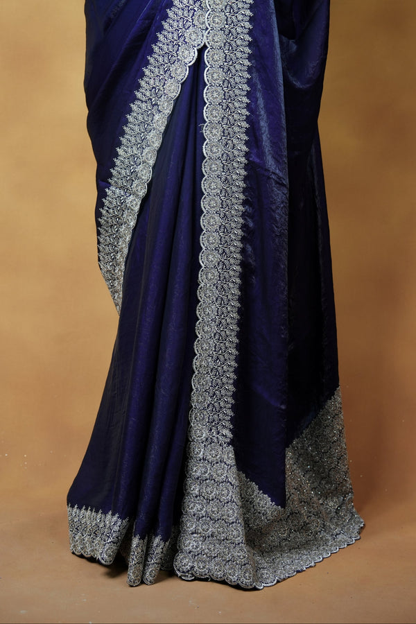 A close-up of a dark blue saree draped elegantly, showcasing its detailed silver-gray border and pallu against a solid warm brown background.