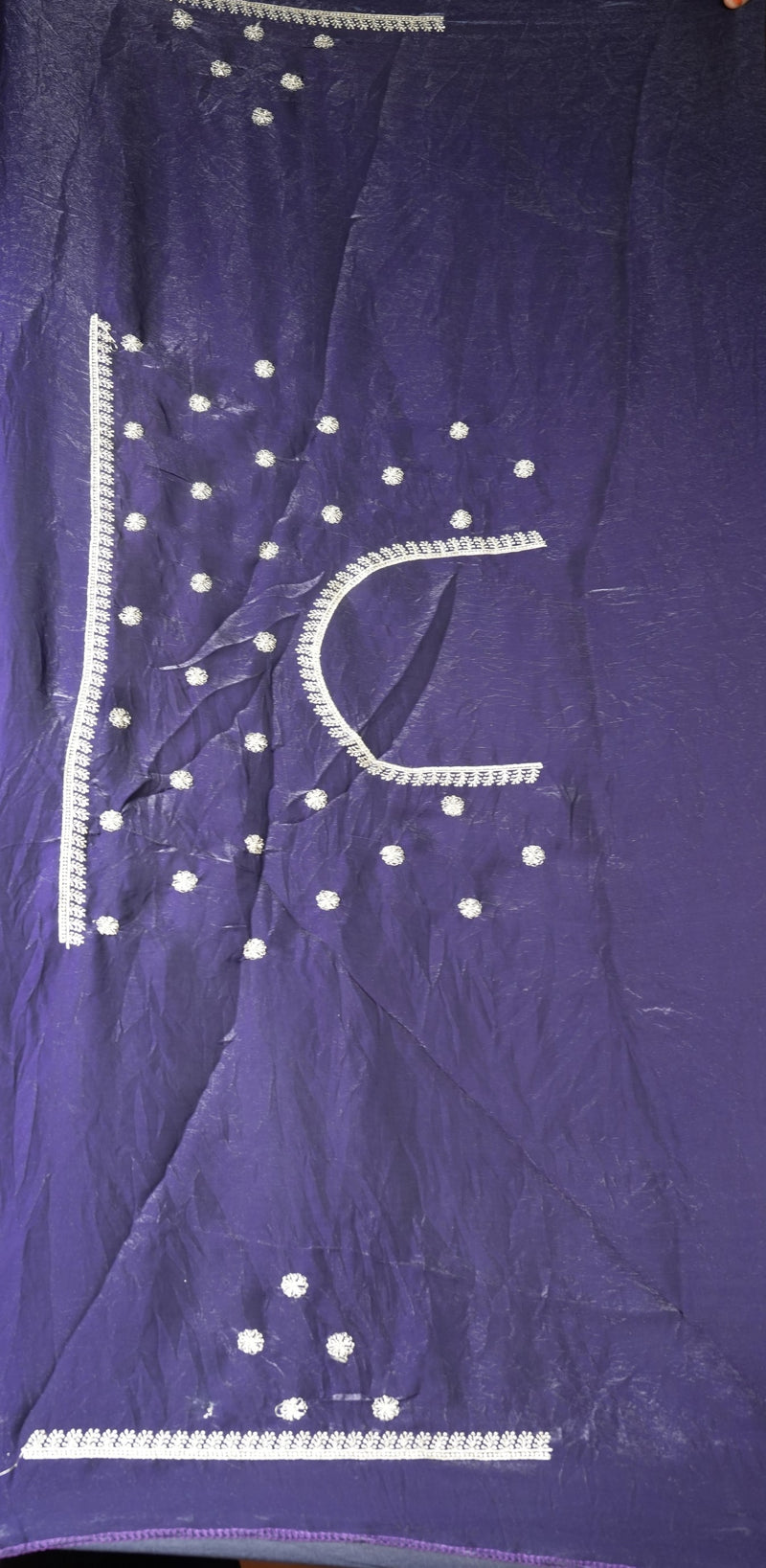 A close-up of a purple fabric, likely a traditional ethnic garment like a kurta or salwar suit material, decorated with white sequins and silver thread embroidery forming a stylized letter 'C' and dotted patterns.