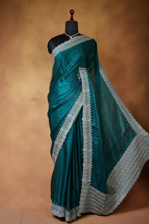 A teal green silk saree with intricate silver and white border embroidery, displayed on a black mannequin torso against a warm brown textured backdrop.