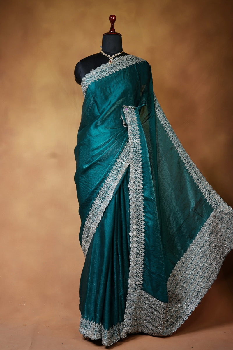 A teal green silk saree with intricate silver and white border embroidery, displayed on a black mannequin torso against a warm brown textured backdrop.