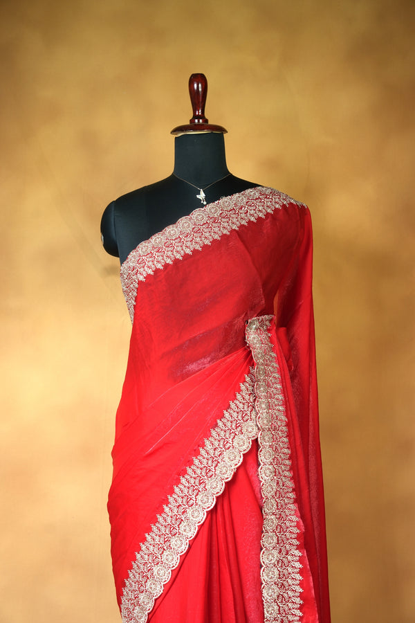 A bright red sari with intricate white and beige floral lace borders, draped on a black mannequin torso against a warm, textured golden-brown studio background.