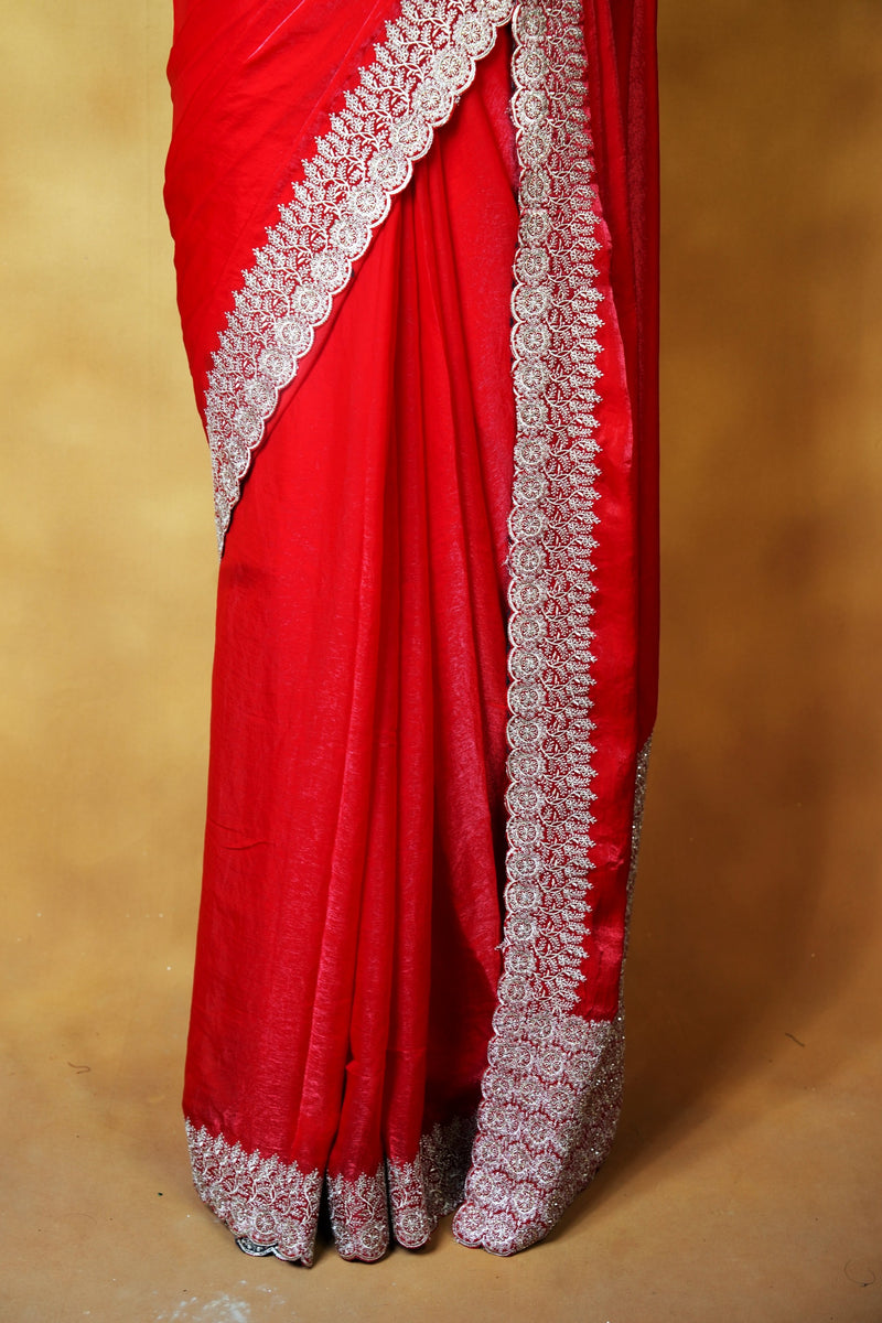 A close-up of a bright red sari with ornate silver and white embroidered borders, draped elegantly against a warm beige background.