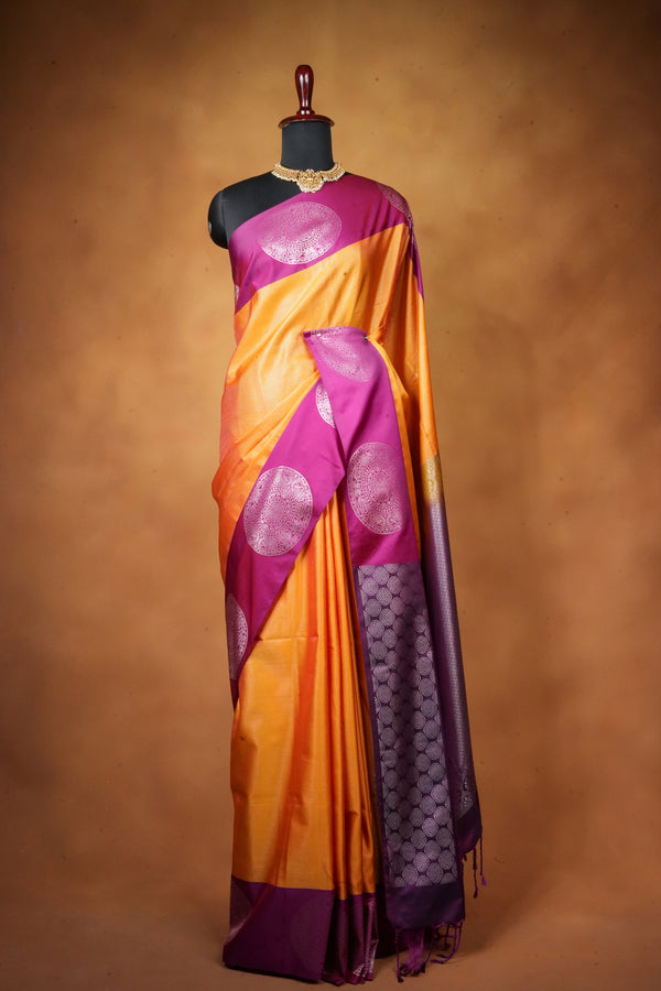 A purple and orange silk saree with metallic circular motifs, draped on a black mannequin torso against a warm brown studio backdrop.
