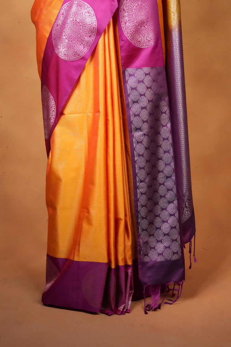 A close-up of a draped silk saree in vibrant orange and purple, featuring intricate metallic zari patterns and a border, set against a solid tan background.