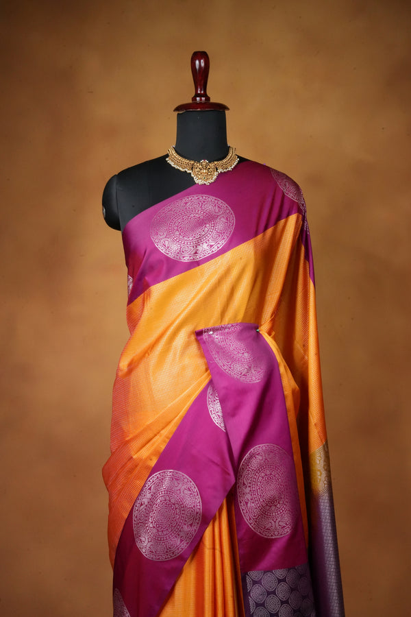 A purple and orange silk saree with metallic circular motifs, draped on a black mannequin torso against a warm brown studio background.