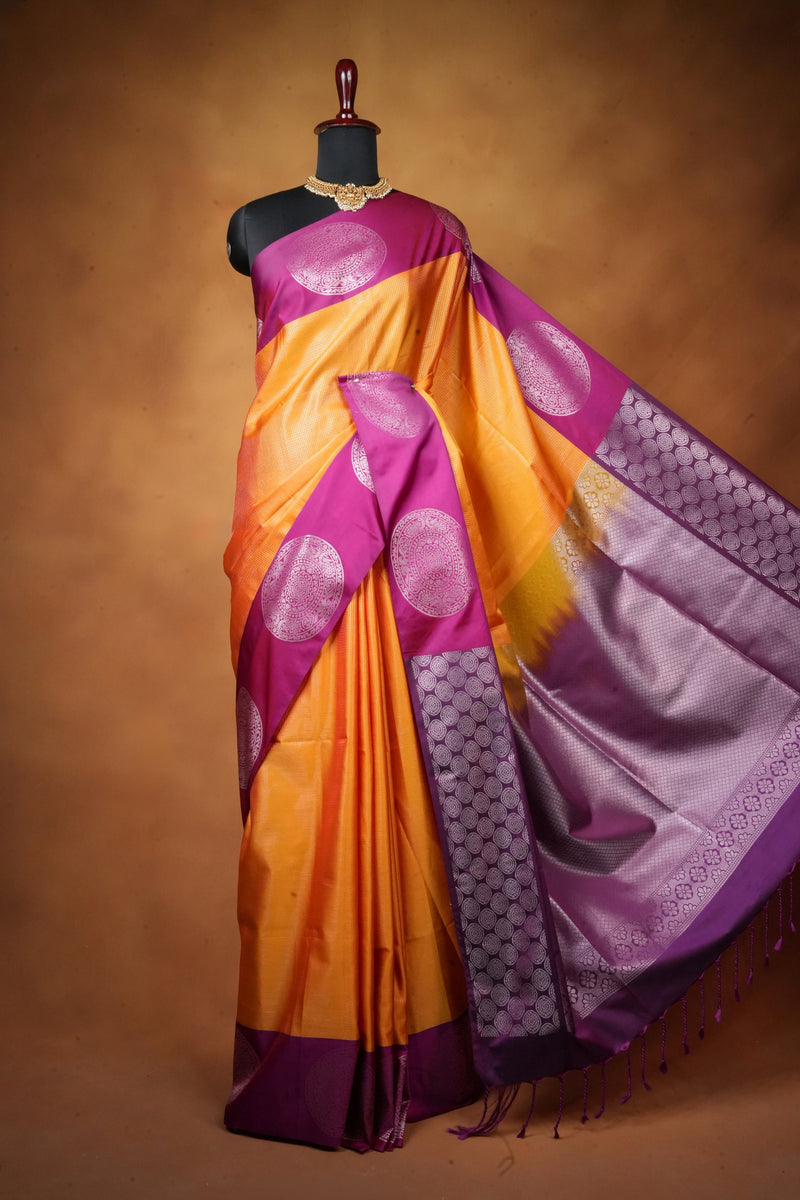 A purple and orange silk saree with metallic silver motifs, displayed on a black mannequin against a warm brown studio background.