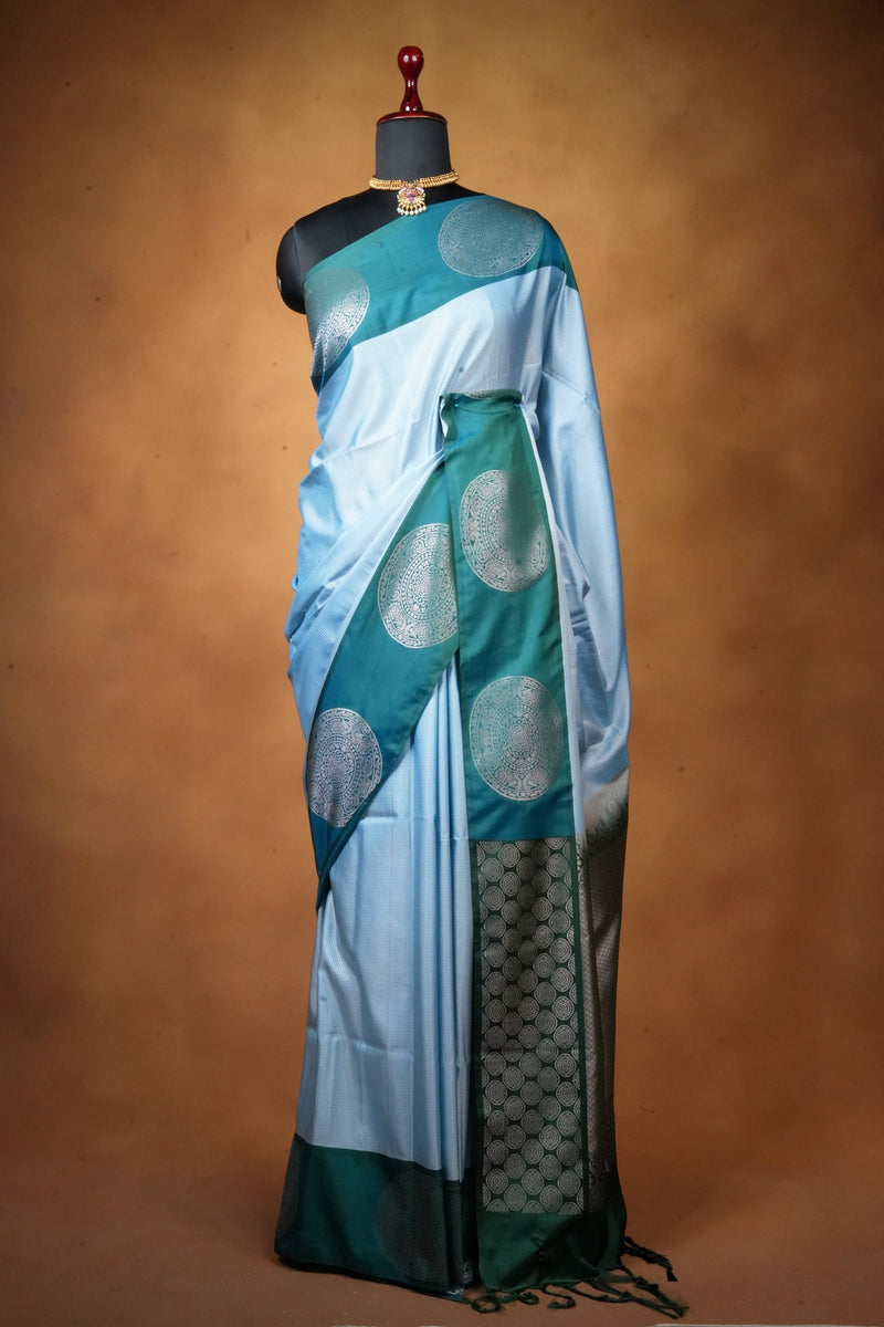 A light blue and teal saree with silver zari circular motifs and a dark green border, displayed on a black mannequin against a warm brown studio backdrop.