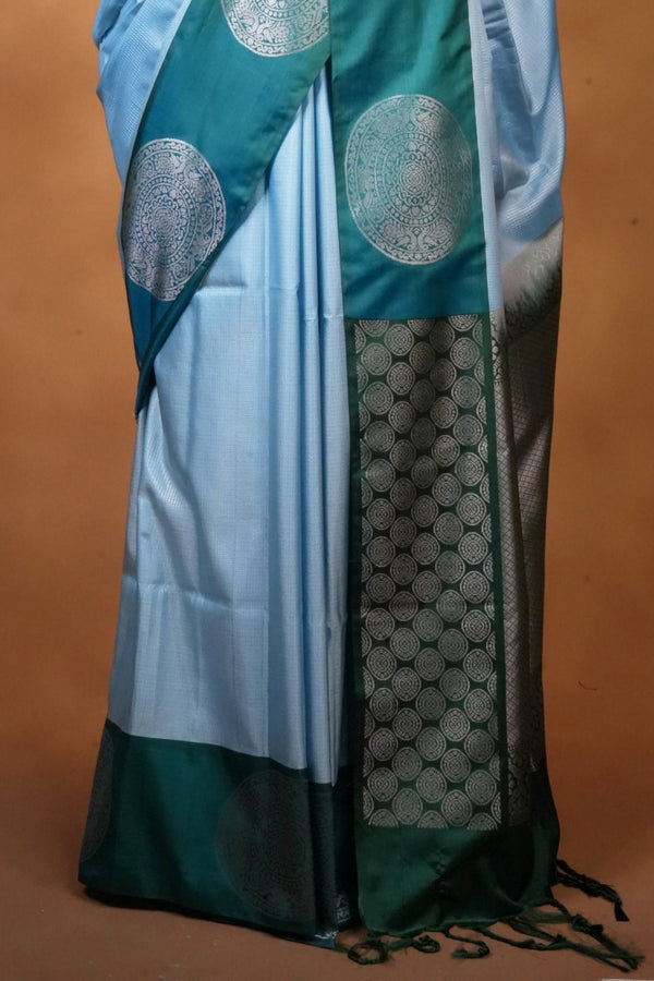 A light blue and teal saree with silver zari circular motifs and a dark green border, displayed on a black mannequin against a warm brown studio backdrop.