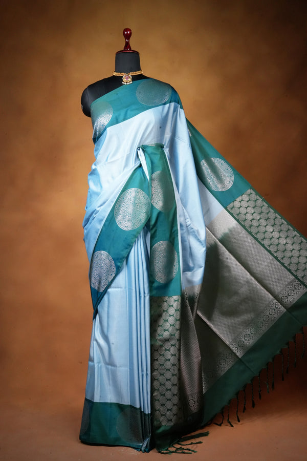 A light blue and teal silk saree with silver zari motifs and a silver border, displayed on a black mannequin against a warm brown studio background.