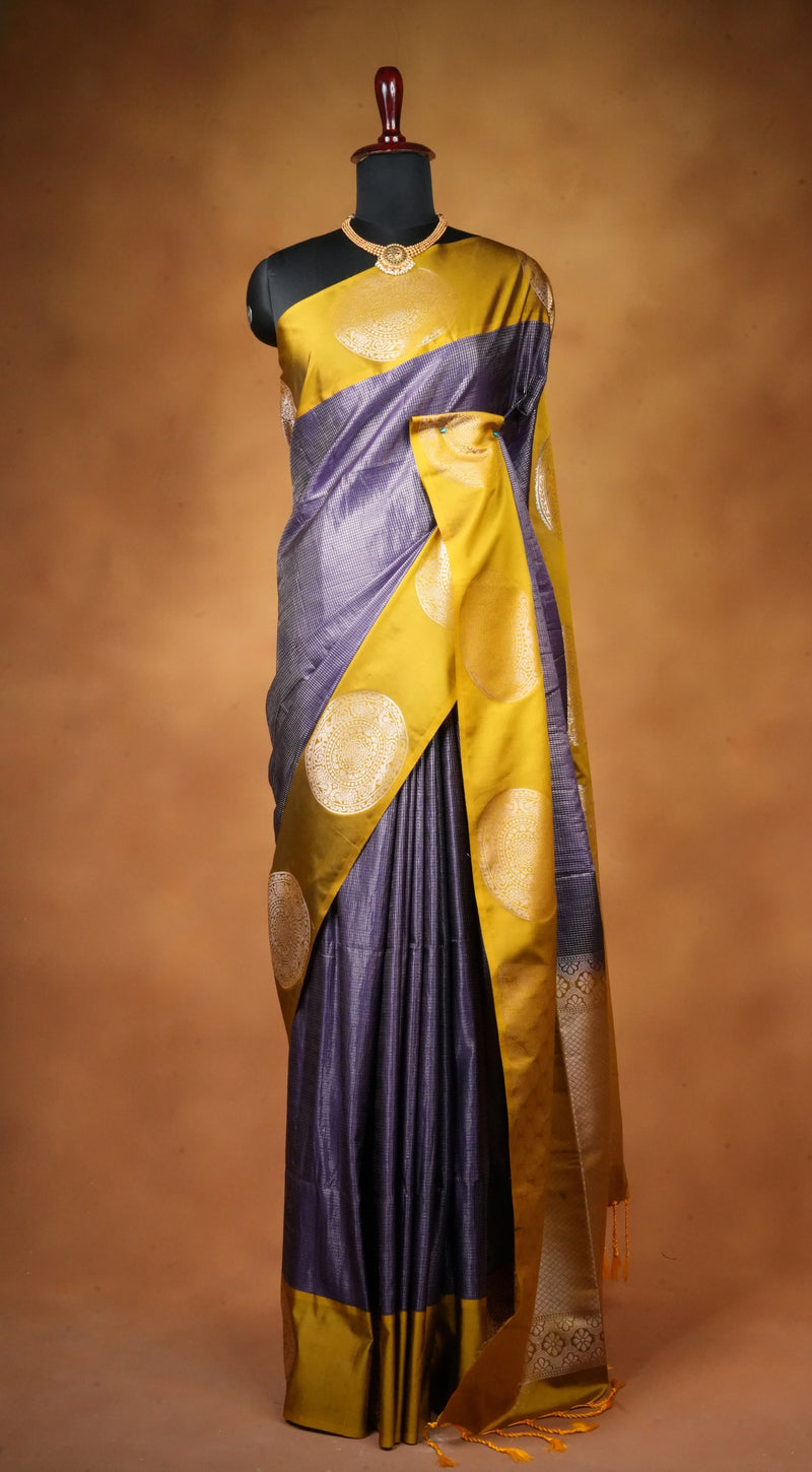 A purple and gold silk saree draped on a black mannequin torso, set against a warm brown studio background. The saree features a purple body with a wide gold border and circular gold zari motifs.