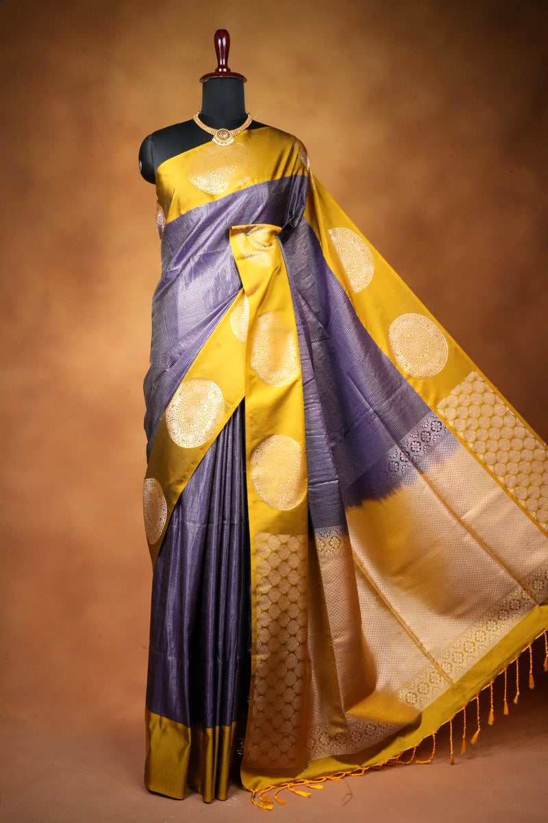 A purple and mustard yellow silk saree with gold zari motifs, draped on a black mannequin against a warm brown studio background.