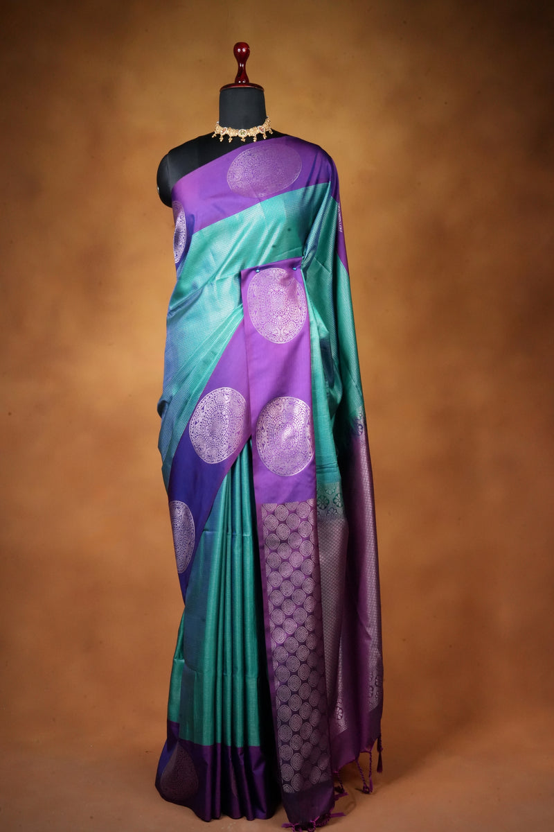 A purple and teal silk saree with metallic circular motifs, draped on a black mannequin torso against a warm brown studio background.