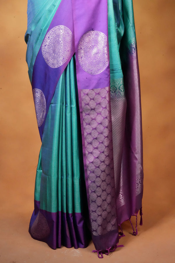 A close-up of a purple and teal silk saree with intricate silver zari motifs, draped against a solid tan background.
