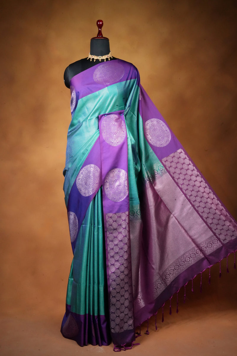 A purple and teal Banarasi silk saree with intricate zari work, draped on a black mannequin against a warm brown studio background.