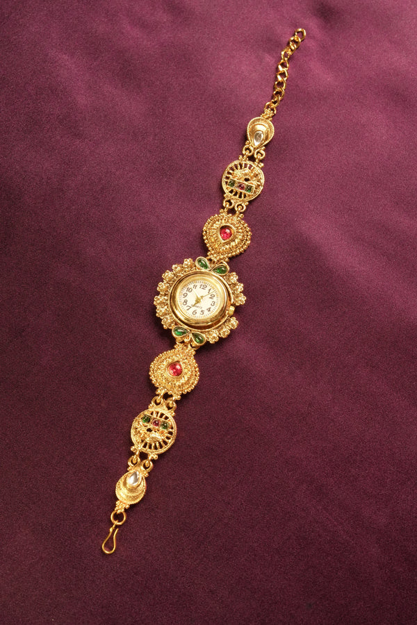 An ornate gold-toned traditional watch bracelet with a central round dial, decorated with red and green stones, set against a dark purple textured background.