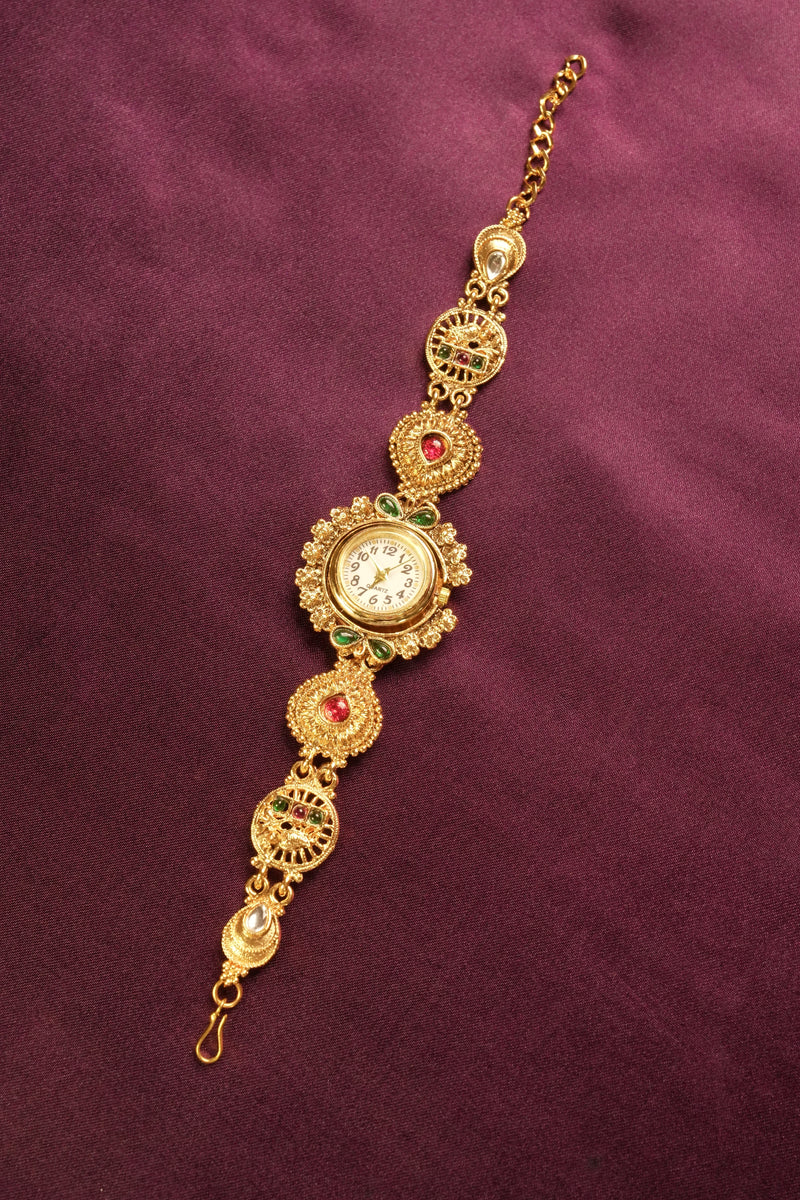An ornate gold-toned traditional watch bracelet with a central round dial, decorated with red and green stones, set against a dark purple textured background.